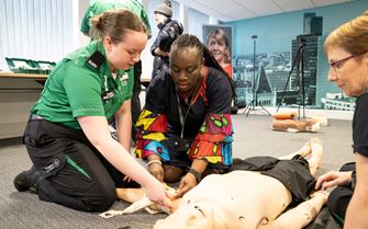 A Cadet First Aider demonstrating to two women how to treat severe bleeding on a mannequin on the floor. A Cadet First Aider demonstrating to two women how to treat severe bleeding on a mannequin on the floor.
